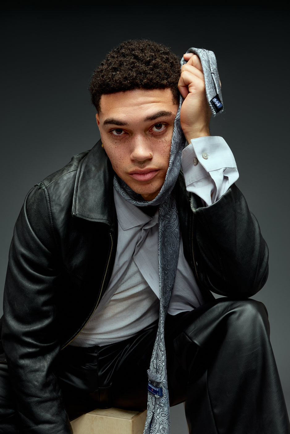 Young man in a black leather jacket, crouched with a hand on his head, editorial studio portrait