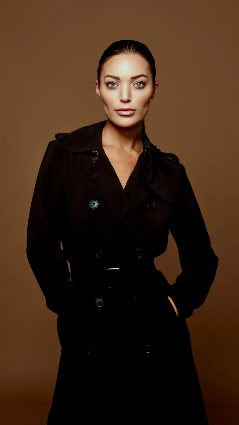 Woman with slicked-back hair in a black trench coat on a warm brown studio backdrop