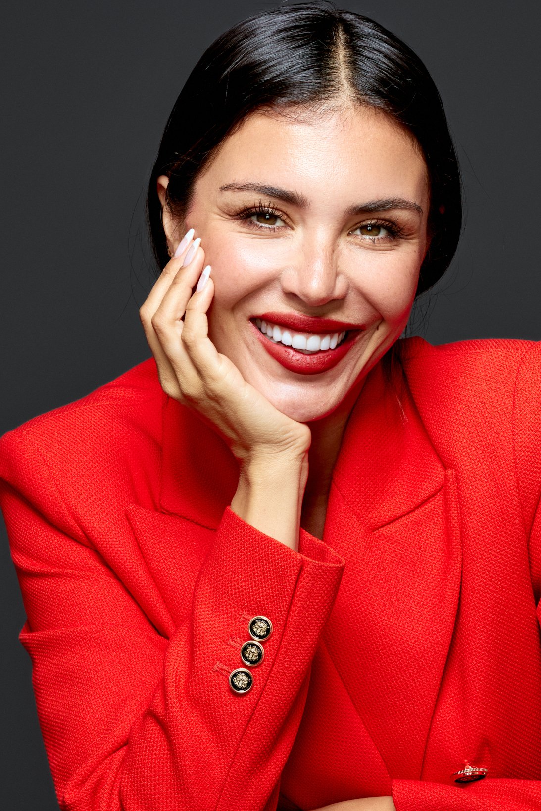 Smiling woman in a red blazer, hand on her cheek, studio portrait